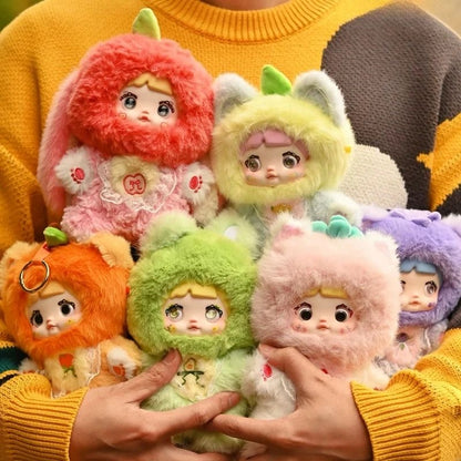 Colorful plush toys with animal-themed hats held by a person wearing a yellow sweater. Nommi V3 Interesting Fruit Dolls Series Dolls plush blind box — all designs lineup (Strawberries, Lemon, Blueberry, Avocado, Orange, Apple, Toffee) — Kuwait — نومي